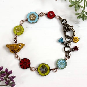 May include: A handcrafted bracelet featuring a charming bird charm and colorful floral beads in shades of red, yellow, blue, and green. The bracelet is strung on copper wire and has a decorative clasp.