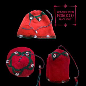Puede incluir: Sombrero rojo de ganchillo con ribetes verdes y blancos y detalles de conchas blancas. El sombrero tiene una borla larga y el texto "100% MADE IN MOROCCO CRAFT SPIRIT" es visible en el fondo.