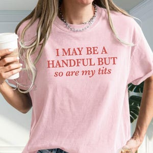 May include: A light pink t-shirt with the text "I MAY BE A HANDFUL BUT so are my tits" in red lettering. The shirt has short sleeves and a crew neck.