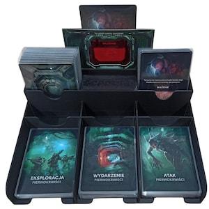 Może przedstawiać: Czarny organizer do gier karcianych z wieloma przegródkami. Organizer mieści stosy kart z grafiką fantasy. Przód organizera ma trzy sloty na karty z polskim tekstem. Góra ma czerwony akcent.