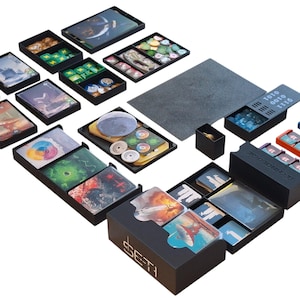 Könnte beinhalten: Eine Sammlung schwarzer Spielboxen und -schalen, gefüllt mit Karten, Spielsteinen und Spielkomponenten. Die Boxen sind auf einer weißen Oberfläche angeordnet. Einige Boxen haben farbenfrohe Kunstwerke. Das Wort "SETI" ist auf einer Box sichtbar.