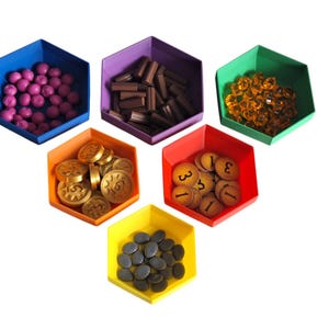 Puede incluir: Componentes de juego de mesa coloridos en recipientes hexagonales. Incluye esferas moradas, bloques marrones, gemas naranjas transparentes, monedas de oro, fichas de madera numeradas y piedras negras. Los recipientes son azules, morados, verdes, naranjas, rojos y amarillos.