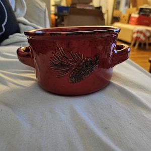 Op de afbeelding: Een rode keramische soepkom met twee handvatten. De kom heeft een ontwerp met een dennenappel en dennennaalden.