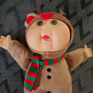 May include: A Cabbage Patch Kid doll dressed in a brown reindeer costume with a red and green striped scarf. The doll has rosy cheeks, open arms, and a smiling expression. The costume includes antlers and a hood.