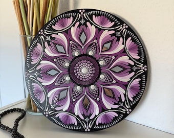 May include: A purple and white mandala painted on a round wooden plaque. The mandala features a repeating pattern of flower-like shapes and dots. The plaque is hanging on a white wall.