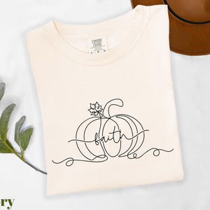 A white t-shirt with a black line drawing of a pumpkin and the word "faith" printed on it, along with a brown hat and some greenery.