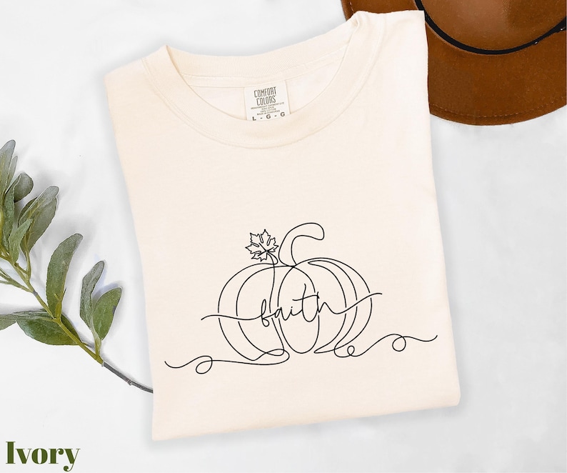 A white t-shirt with a black line drawing of a pumpkin and the word "faith" printed on it, along with a brown hat and some greenery.