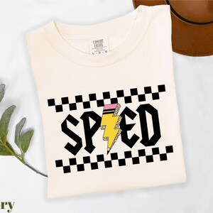 a white t-shirt with a graphic design on it, including a lightning bolt and checkered patterns. The t-shirt is placed on a white surface, with a brown hat and a green plant visible in the background.
