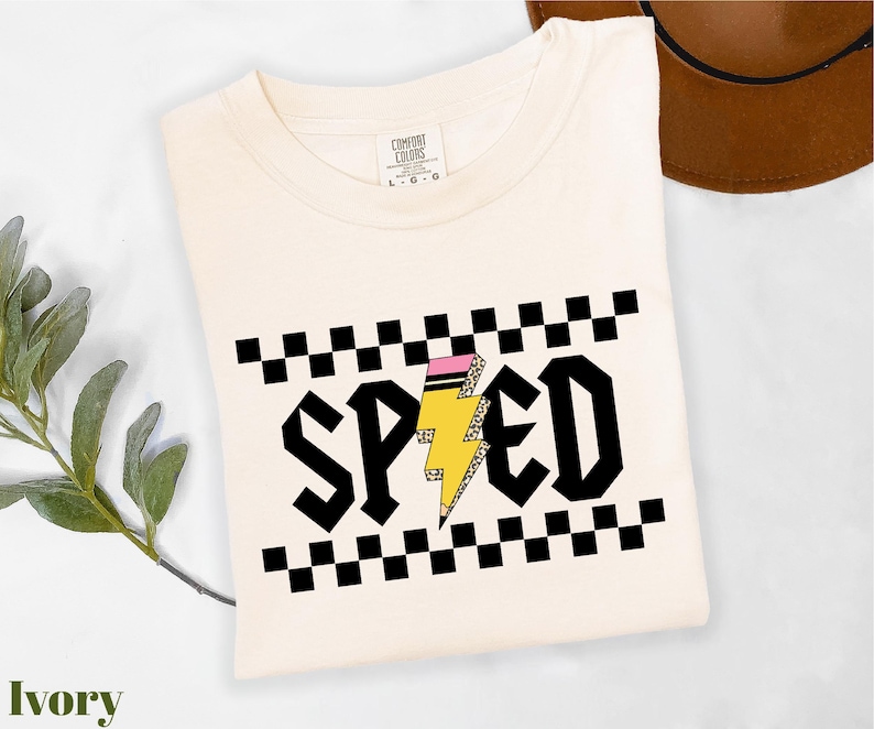 a white t-shirt with a graphic design on it, including a lightning bolt and checkered patterns. The t-shirt is placed on a white surface, with a brown hat and a green plant visible in the background.