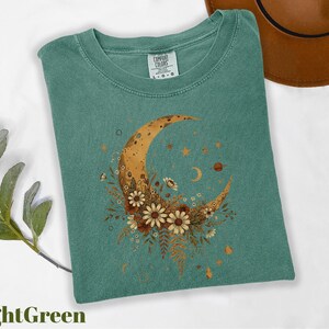 a green t-shirt with a crescent moon and floral design, along with a brown hat and some greenery.