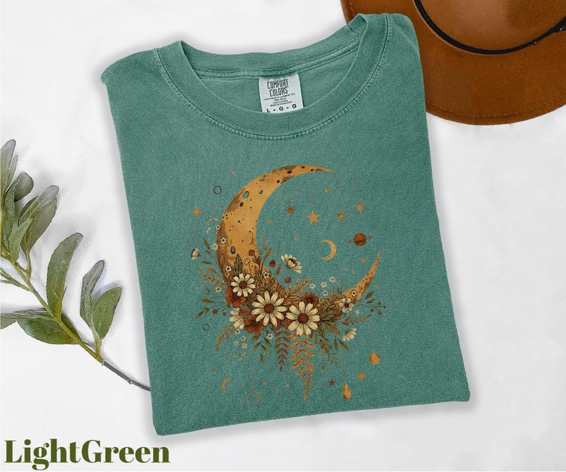 a green t-shirt with a crescent moon and floral design, along with a brown hat and some greenery.