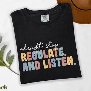 May include: Black t-shirt with the words "alright stop. REGULATE. AND LISTEN." in colorful, rounded letters. The shirt is folded and has a tag that says "Comfort Colors". A brown hat is in the background.
