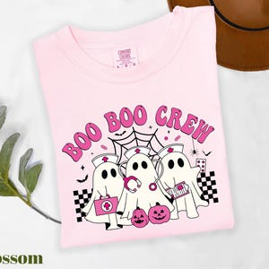 May include: A light pink t-shirt with the words "BOO BOO CREW" in pink lettering. The graphic features three ghost characters dressed as nurses, a spider web, bats, and pumpkins. The shirt is labeled "Blossom".