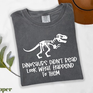 May include: A dark gray t-shirt with a white dinosaur skeleton graphic and the text "Dinosaurs Didn't Read Look What Happened To Them." The shirt is a Comfort Colors brand.