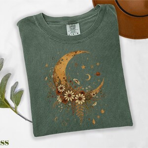a green t-shirt with a moon and star design, along with some flowers and leaves, placed on a white surface. There is also a brown hat visible in the background.