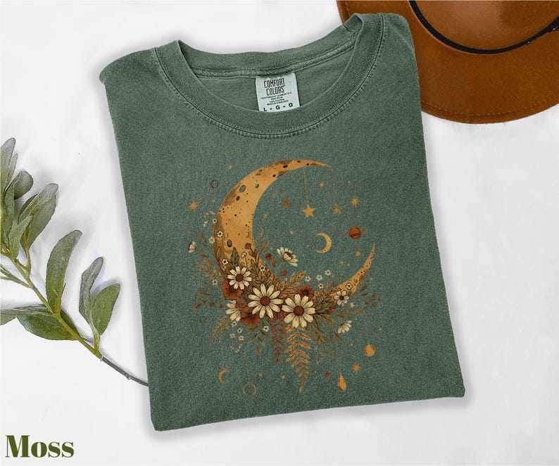 a green t-shirt with a moon and star design, along with some flowers and leaves, placed on a white surface. There is also a brown hat visible in the background.