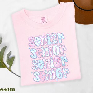 A pink t-shirt with a colorful text design that says "senior senior senior" printed on it, along with some decorative elements. The t-shirt is placed on a white surface, and there is a brown hat visible in the background.