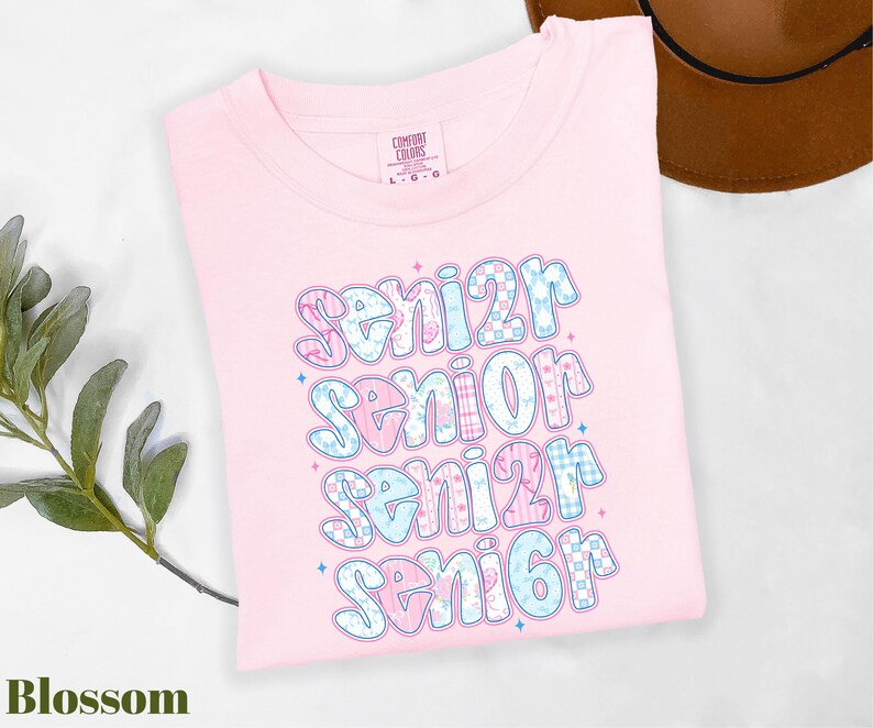 A pink t-shirt with a colorful text design that says "senior senior senior" printed on it, along with some decorative elements. The t-shirt is placed on a white surface, and there is a brown hat visible in the background.