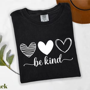 A black t-shirt with a white heart and the phrase "be kind" printed on it, along with a brown hat and some greenery.