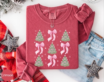 May include: Crimson long-sleeve shirt with a crew neck. The shirt features a design of green Christmas trees and pink bows. A silver necklace with a sun pendant is on the shirt. The shirt is folded with a pair of blue jeans and a star-shaped decoration.