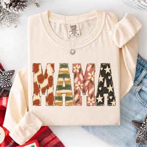 May include: An ivory long-sleeve t-shirt featuring the word MAMA in large block letters. Each letter has a distinct pattern: brown cow print, green and gold stripes, pink flowers, and black with white stars and hearts. A tag reads Comfort Colors.