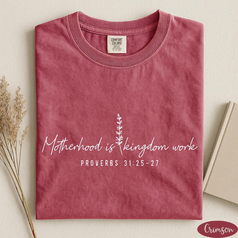 A red t-shirt with white text and a small plant graphic, along with a book and some dried flowers, arranged on a white surface.
