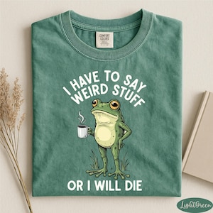 a green t-shirt with a cartoon frog holding a cup of coffee and the text "I HAVE TO SAY WEIRD STUFF OR I WILL DIE" printed on it.