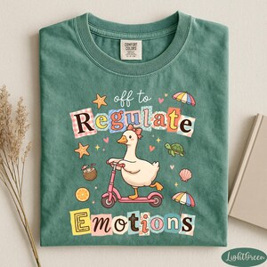 A green t-shirt with a colorful graphic design featuring a duck riding a scooter, surrounded by various objects such as umbrellas, flowers, and a book.