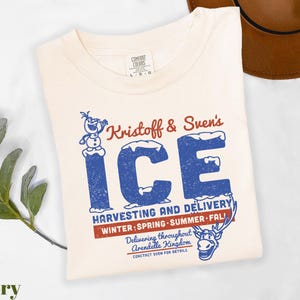 May include: Ivory-colored t-shirt featuring the text "Kristoff & Sven's ICE" in blue and red, with illustrations of Olaf and Sven. The shirt also includes the text "Harvesting and Delivery" and "Winter, Spring, Summer, Fall."