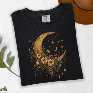 A black t-shirt with a floral design featuring a crescent moon, stars, and flowers, along with a brown hat and some greenery.