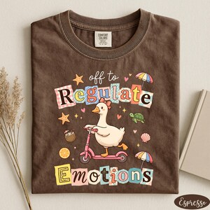 a brown t-shirt with a colorful graphic design depicting a duck riding a pink scooter, surrounded by various objects such as a star, umbrella, and seashell.