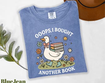 May include: Blue jean colored t-shirt with a graphic of a duck carrying books, surrounded by flowers and the text "OOOPS, I BOUGHT ANOTHER BOOK". A brown hat is in the background.