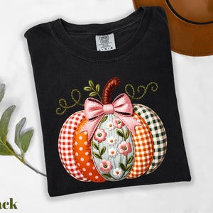 A black t-shirt with a colorful pumpkin embroidered on it, along with a brown hat and some greenery, all placed on a white surface.
