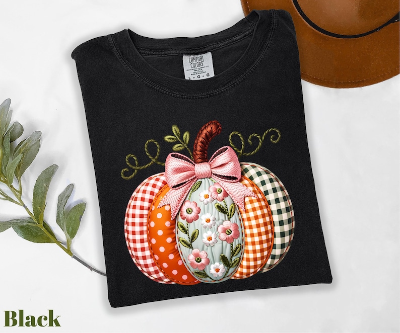 A black t-shirt with a colorful pumpkin embroidered on it, along with a brown hat and some greenery, all placed on a white surface.