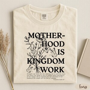 a beige t-shirt with a black and white graphic design that reads "MOTHER-HOOD IS KINGDOM WORK" and includes a quote from the Bible verse Proverbs 3:25-27.