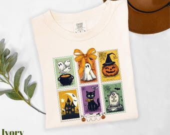 May include: Ivory-colored t-shirt featuring a colorful Halloween-themed stamp design. The stamps depict a cauldron, ghost, pumpkin, haunted house, black cat, and tombstone. The shirt is a Comfort Colors L-G-G.