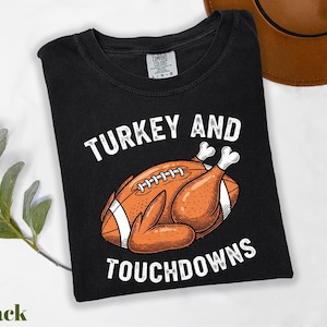 May include: Black t-shirt with a graphic of a football-turkey hybrid and the words "TURKEY AND TOUCHDOWNS" in white. The design features an orange football with turkey legs. A brown hat is visible in the background.