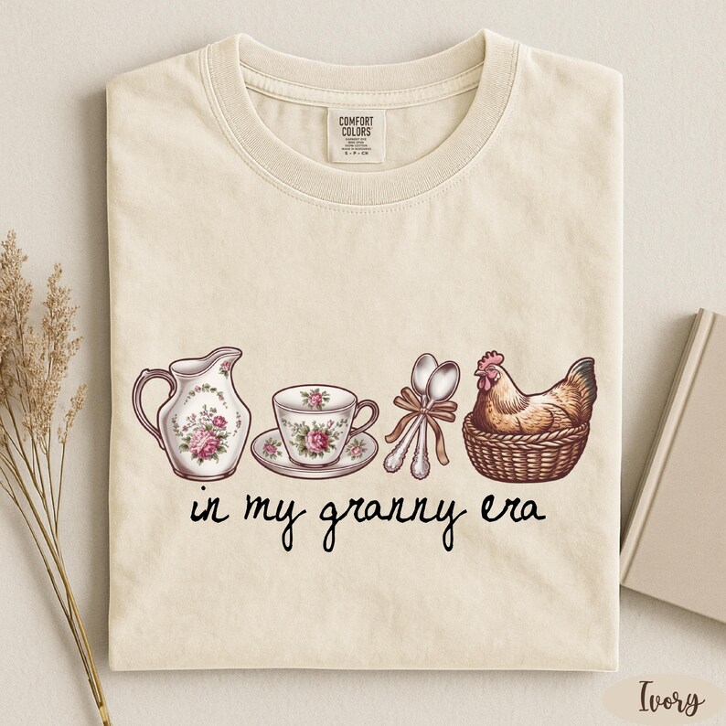 A beige t-shirt with a printed design depicting a tea set, a rooster, and a basket, along with the text "in my granny era" printed on it.