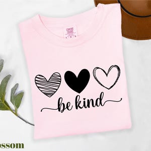 A pink t-shirt with the text "be kind" printed on it, along with three heart symbols. The t-shirt is placed on a white surface, and there is a brown hat visible in the background.