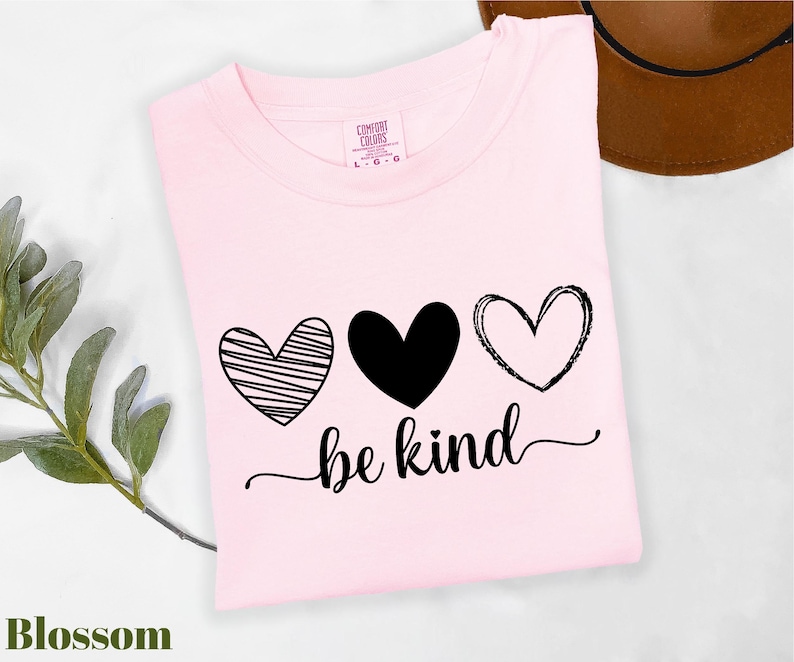 A pink t-shirt with the text "be kind" printed on it, along with three heart symbols. The t-shirt is placed on a white surface, and there is a brown hat visible in the background.