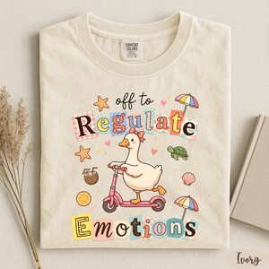 a t-shirt with a colorful and whimsical design, including a duck riding a scooter, surrounded by various objects such as umbrellas, flowers, and a book.