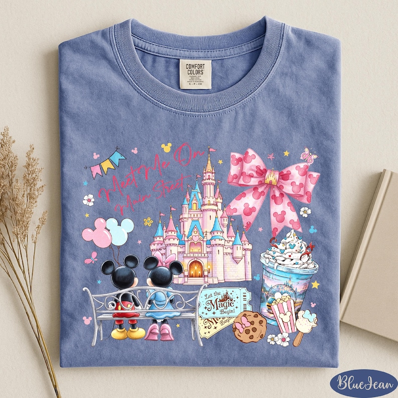 A blue t-shirt with a Disney-themed design, including Mickey Mouse, a castle, and various Disney-related items such as a cup of ice cream and a cookie.