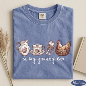 A blue t-shirt with a printed design of a tea set, a chicken in a basket, and the text "in my granny era" printed on it.
