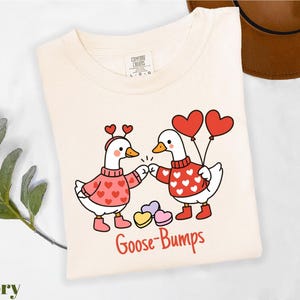 A white t-shirt with a graphic design featuring two cartoon geese wearing heart-patterned sweaters and holding heart-shaped balloons. The text "Goosebumps" is written below the image.