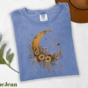 A blue denim t-shirt with a crescent moon and floral design is displayed on a white surface, accompanied by a brown hat and a green plant.