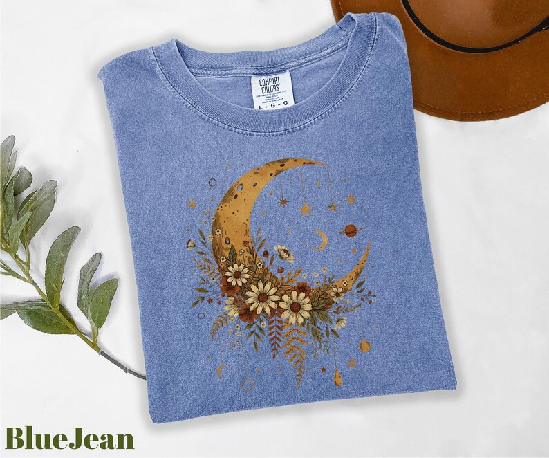 A blue denim t-shirt with a crescent moon and floral design is displayed on a white surface, accompanied by a brown hat and a green plant.