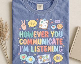 Può includere: Una t-shirt blu a girocollo con una grafica colorata e il testo "HOWEVER YOU COMMUNICATE I'M LISTENING". Il design include illustrazioni di un segno di pace, fumetti, una cartellina, un tablet con icone e delle stelle.