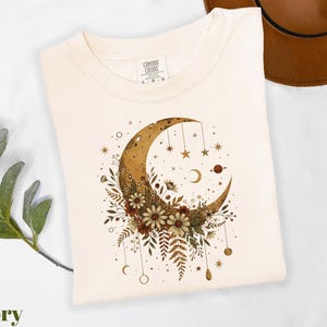 A white t-shirt with a crescent moon and floral design printed on it, along with some greenery and a brown hat.