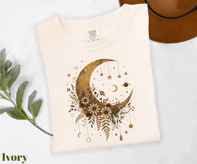 A white t-shirt with a crescent moon and floral design printed on it, along with some greenery and a brown hat.