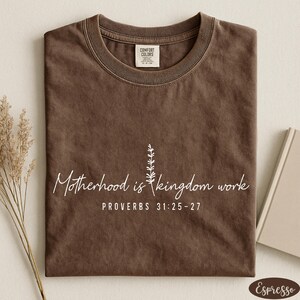 A brown t-shirt with a printed message about motherhood and kingdom work, along with a small plant and a book.